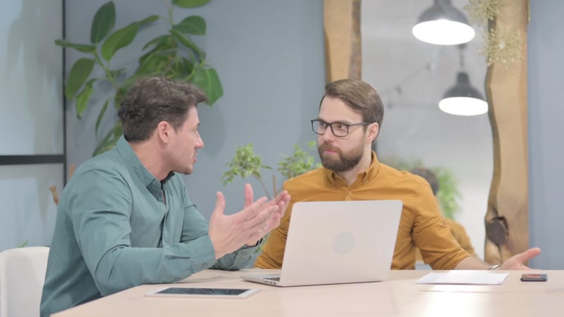 Two men sitting at a desk discussing work with a laptop in front.