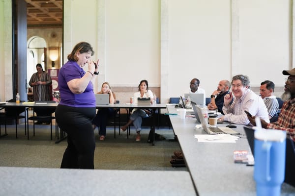 Woman in purple shirt speaking to seated group at conference table.